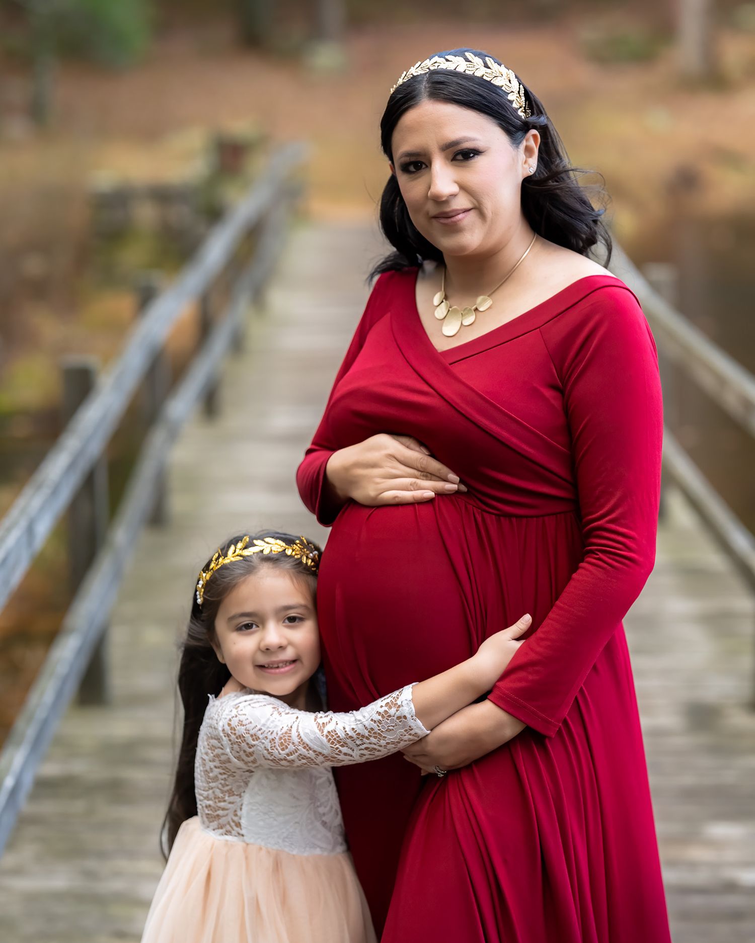 Pregnant woman in red dress poses with young daughter on wooden bridge for maternity photo.