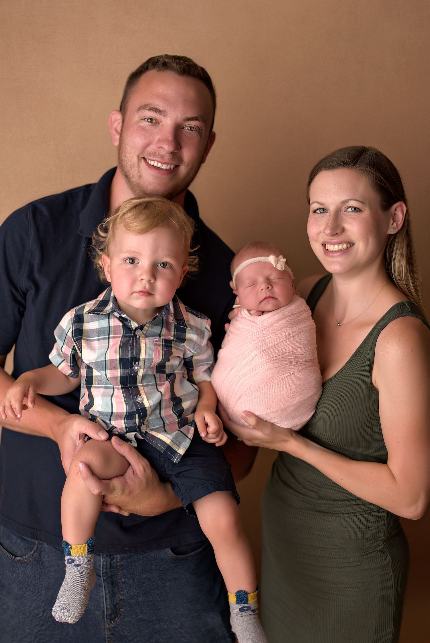 Happy family of four posing for newborn portrait with toddler son and swaddled baby girl in pink.