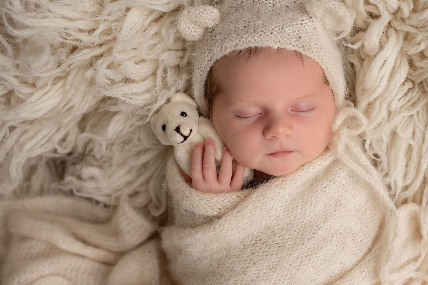 Sleeping newborn baby wrapped in cream knit swaddle, wearing bear bonnet, holding small teddy bear on fluffy white blanket.
