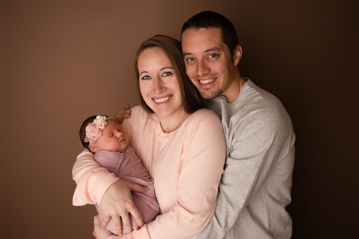 Smiling couple posing with their newborn baby girl wearing a pink floral headband in a professional portrait.