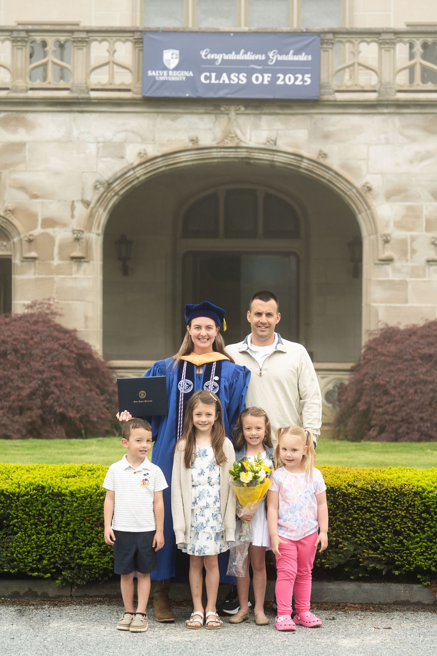 Family celebrates Class of 2025 graduate in blue cap and gown holding diploma outside university building.