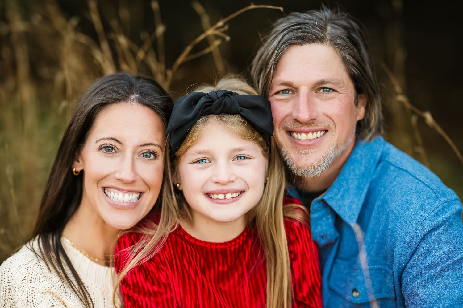 A smiling family of three poses outdoors, featuring a woman, young girl in red, and man in blue shirt.
