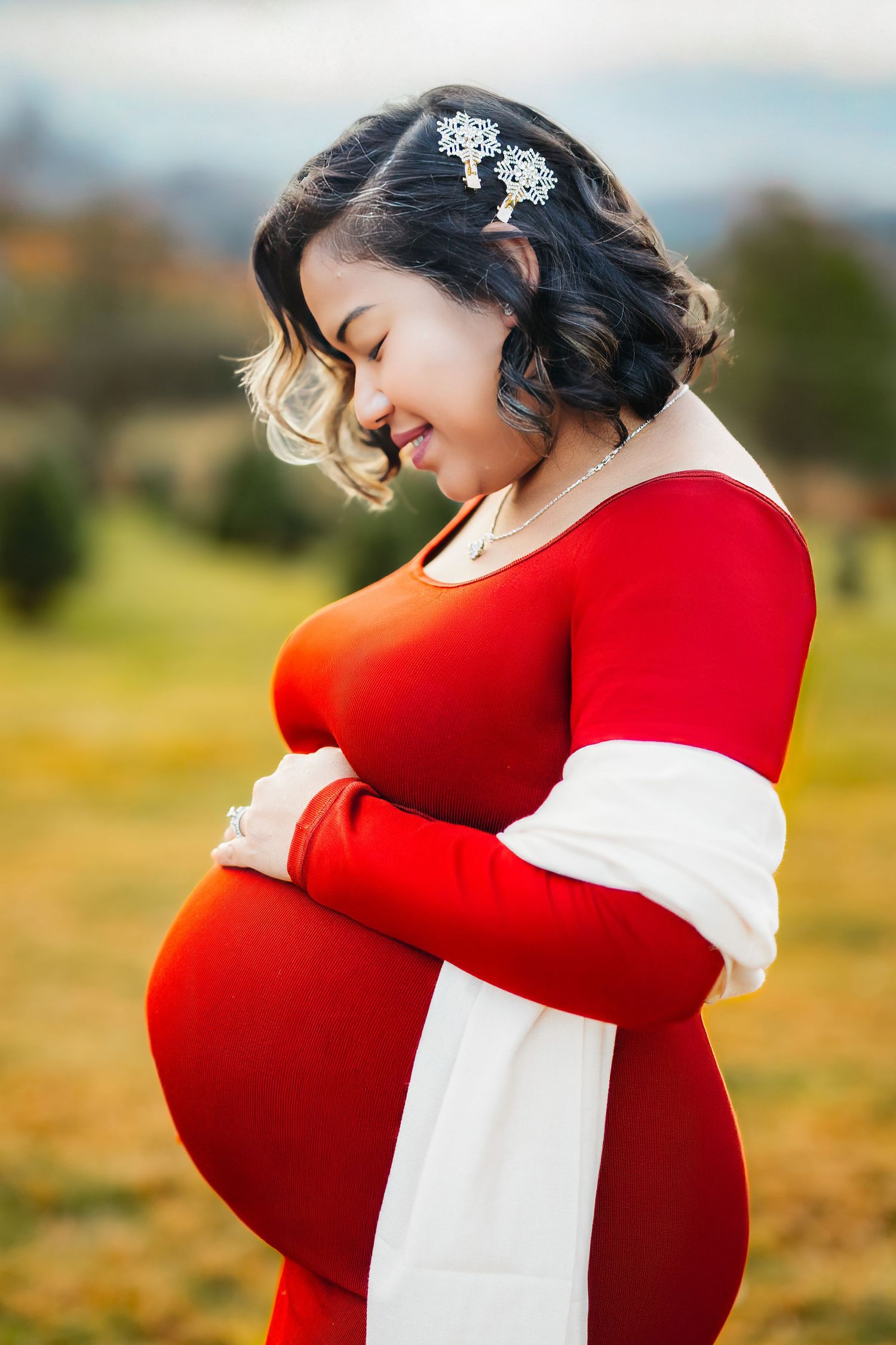 Smiling pregnant woman in red dress holding belly outdoors in a scenic field during autumn.