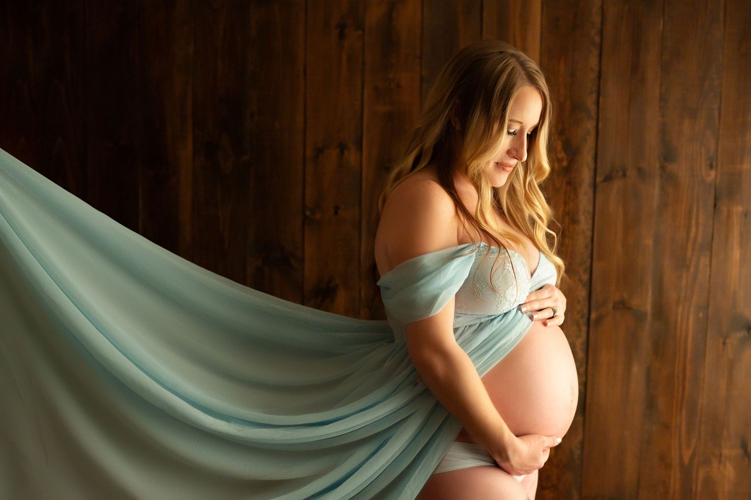 Pregnant woman in flowing mint green fabric wrap poses gracefully against rustic wood wall in maternity photo session.