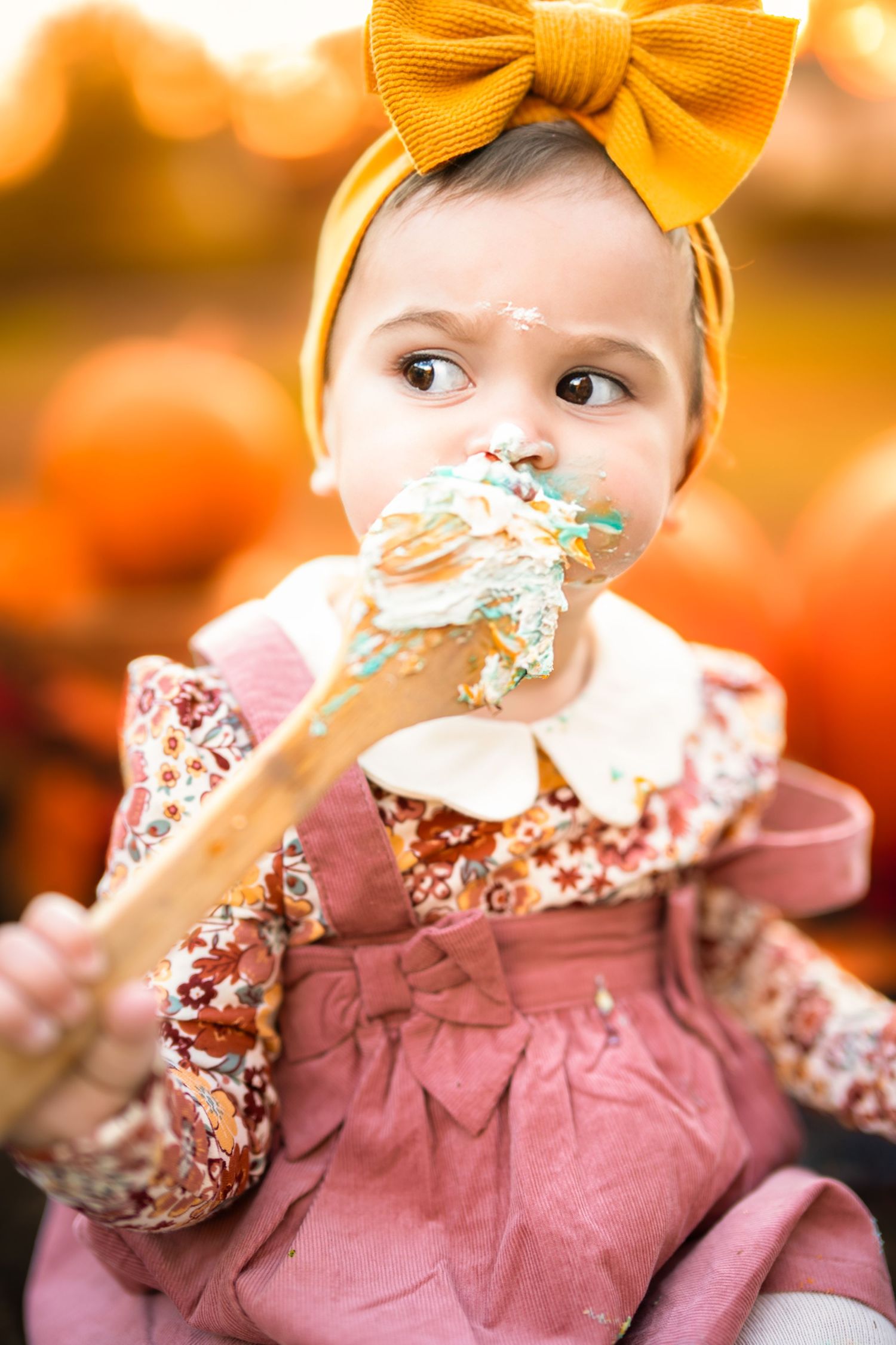 Baby girl in floral dress and mustard bow headband eating cake frosting off a wooden spoon at a pumpkin patch.