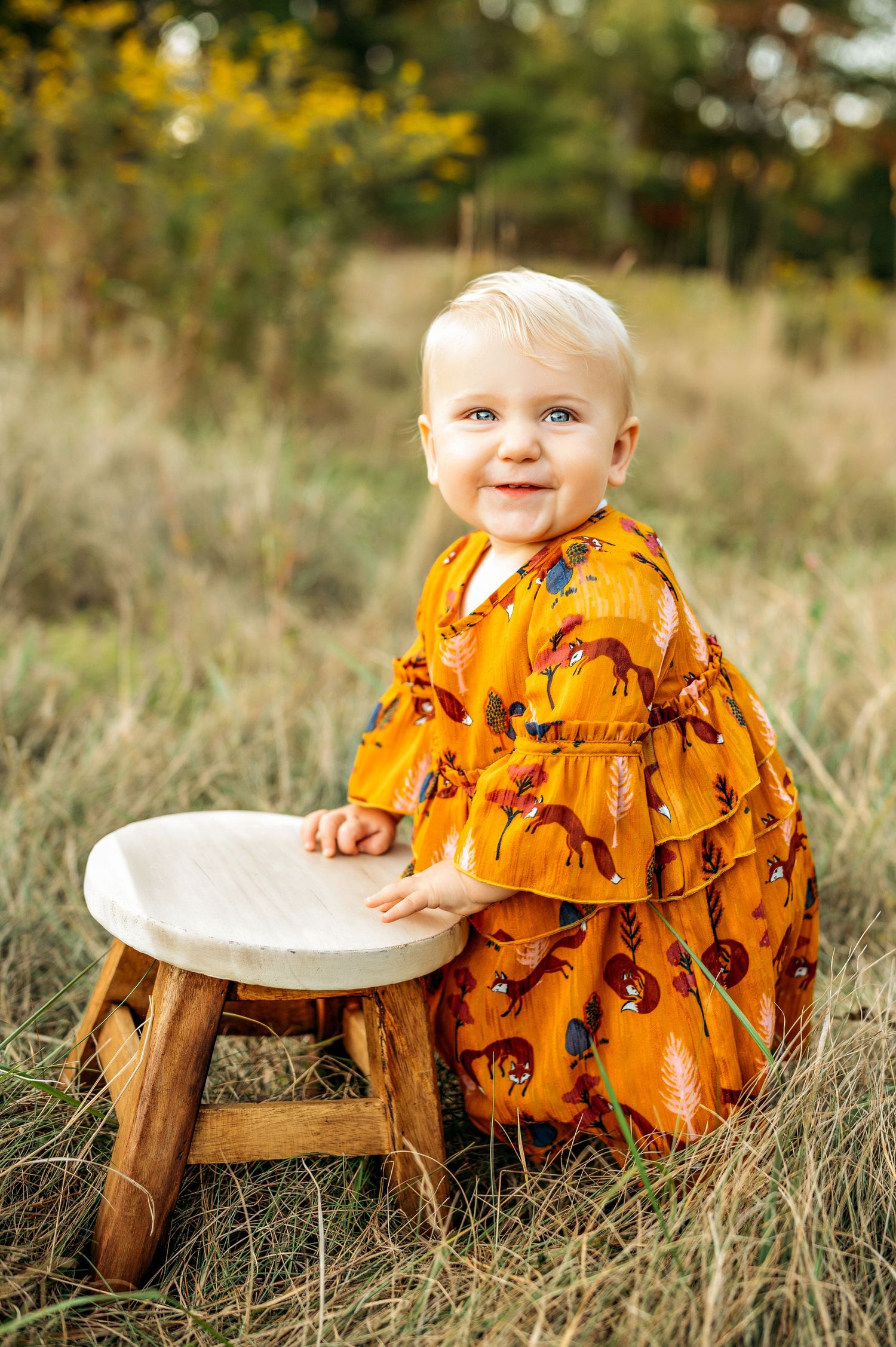 A smiling baby in an orange floral dress stands by a wooden stool in a grassy autumn field.