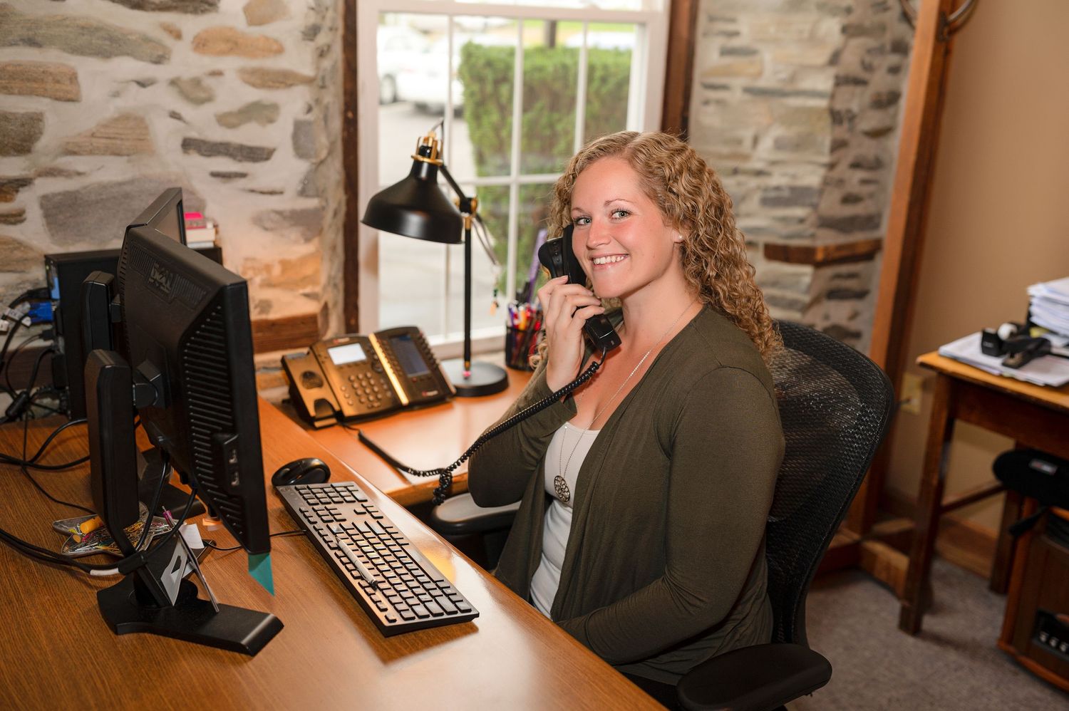 Smiling woman at office desk talking on phone with computer keyboard and lamp in stone wall office.