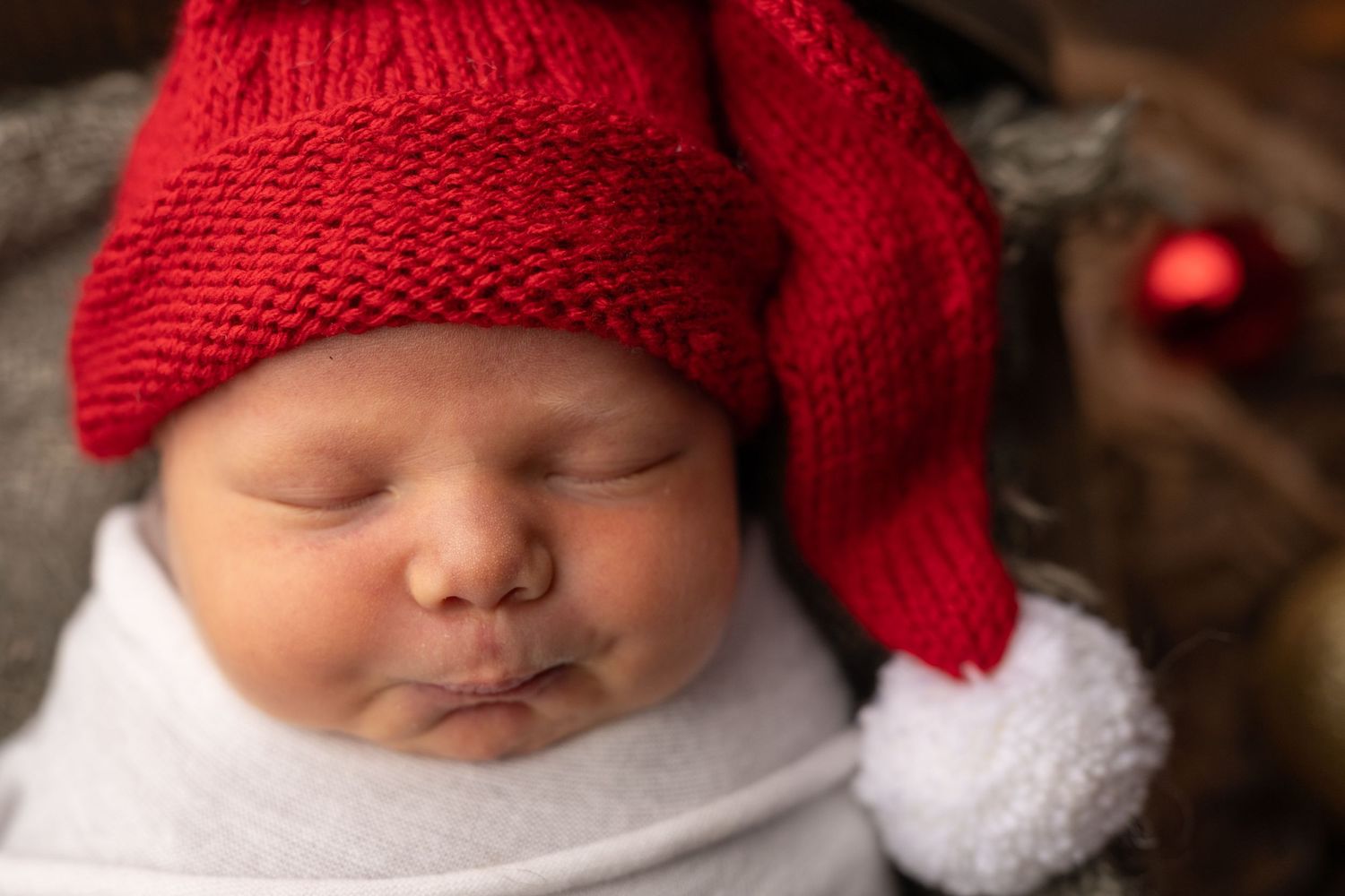 Sleeping newborn baby wearing a red Santa Claus knit hat with white pompom, wrapped in white swaddle for Christmas.