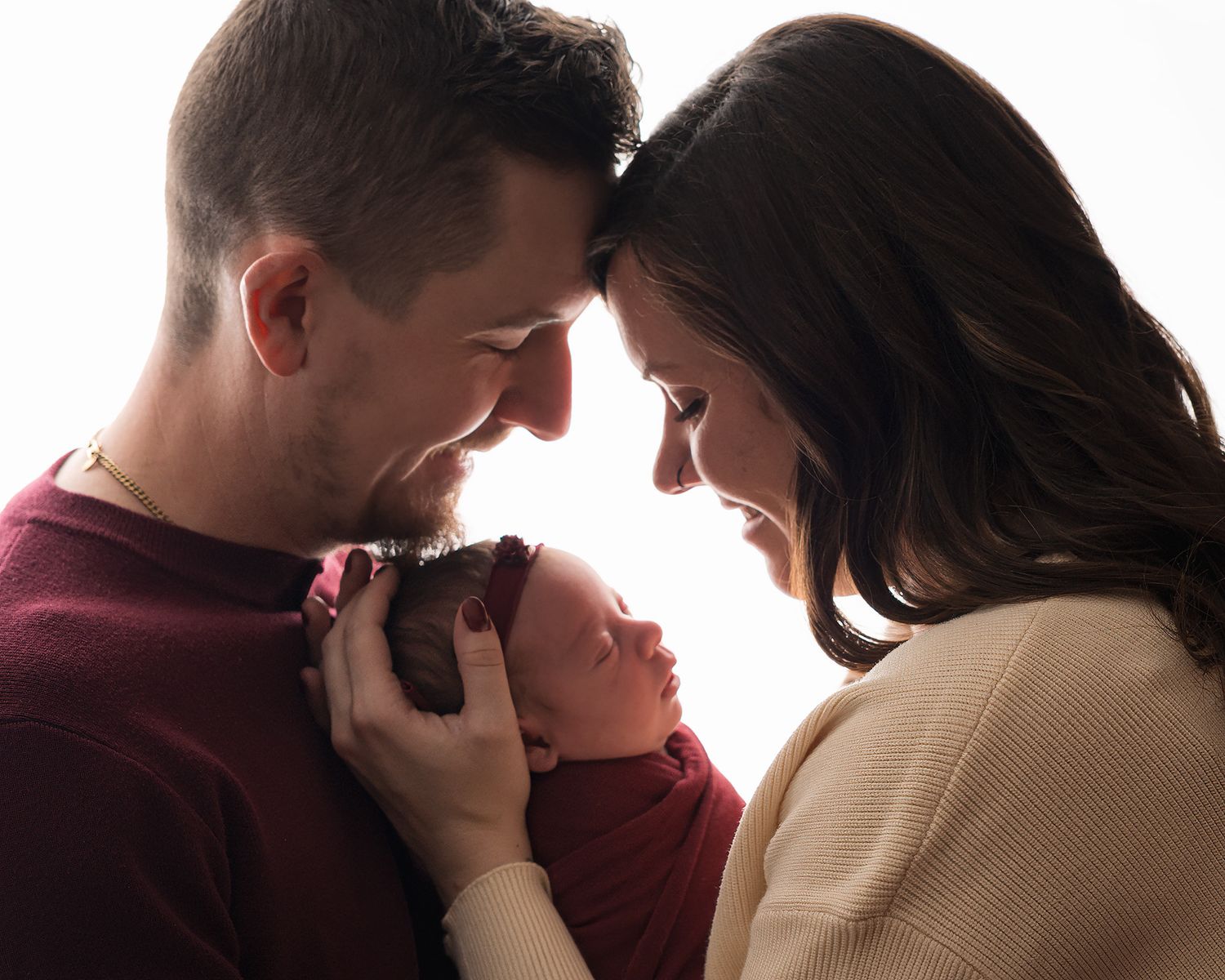 New parents tenderly hold their swaddled newborn baby together, touching foreheads in an intimate studio portrait.