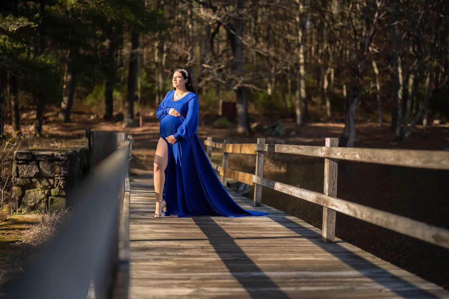 Pregnant woman in elegant royal blue maternity gown posing on wooden bridge surrounded by autumn forest.