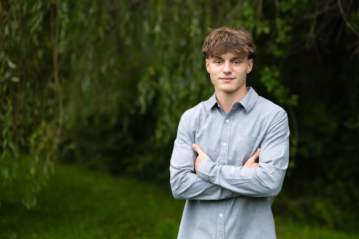 Young man in gray button-down shirt standing with arms crossed in lush green outdoor setting.