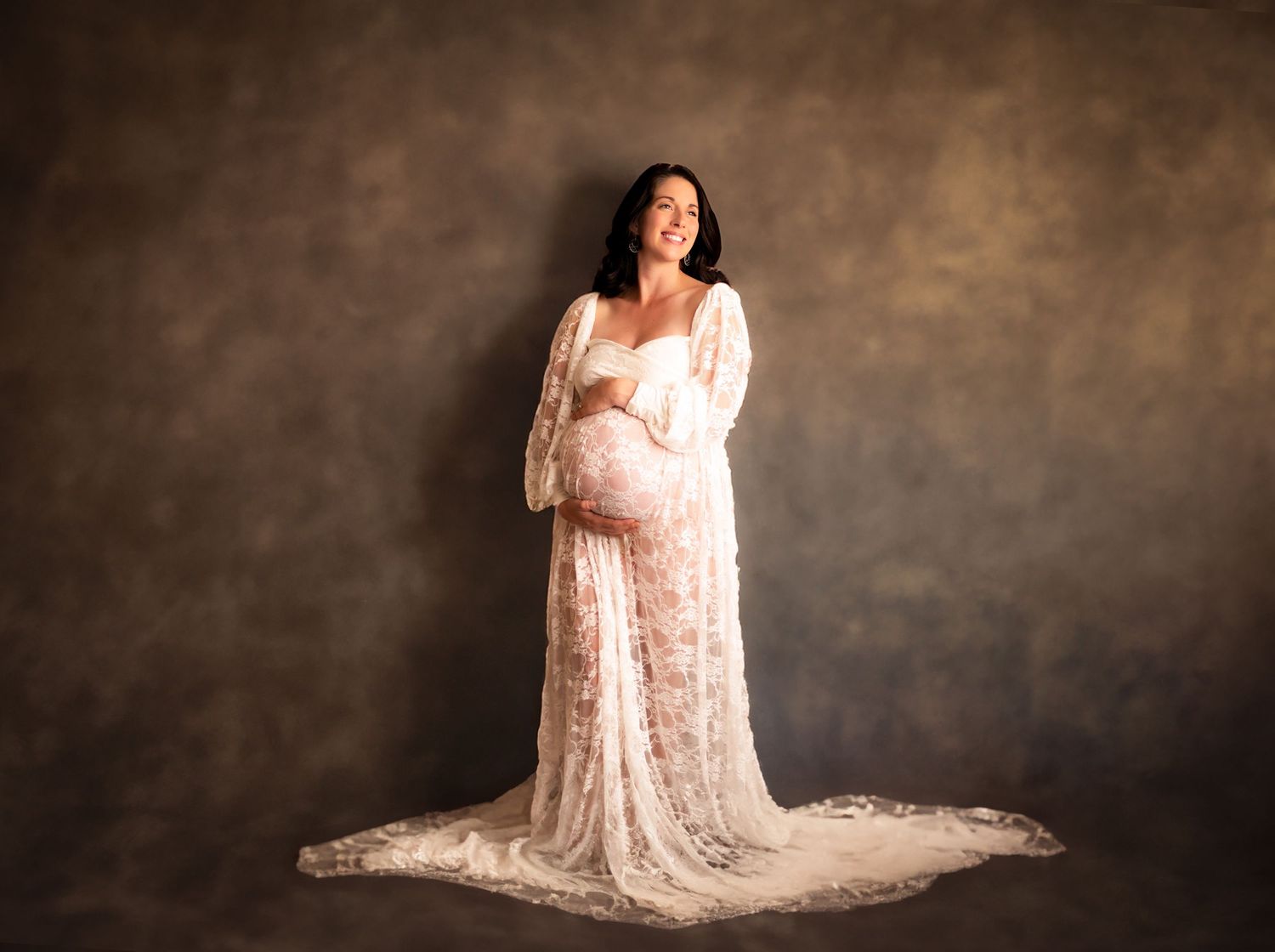 Pregnant woman in elegant white lace maternity gown posing gracefully in a professional studio portrait.