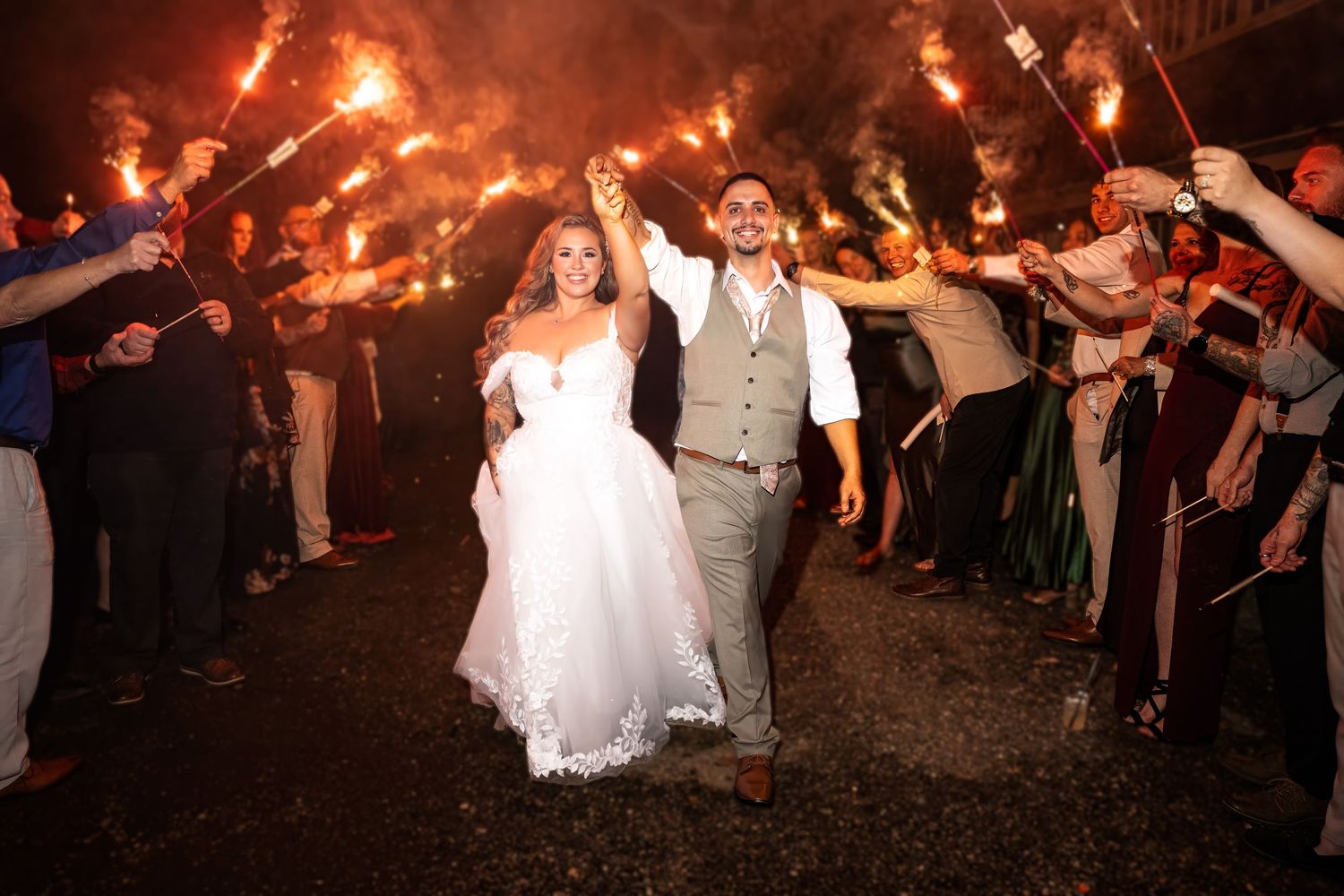 Bride and groom exit through a sparkler tunnel at night, surrounded by cheering wedding guests.