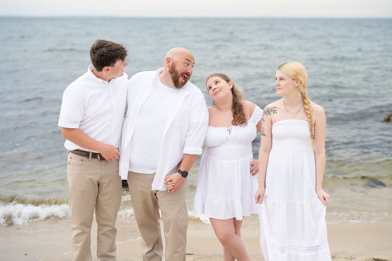 Family of four laughing together on a sandy beach, dressed in coordinating white and khaki outfits.