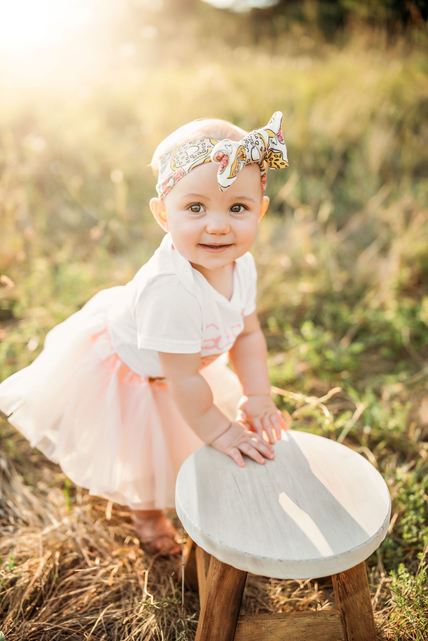 Baby girl in pink tutu and unicorn headband smiles outdoors in golden sunlight, leaning on a small wooden stool.