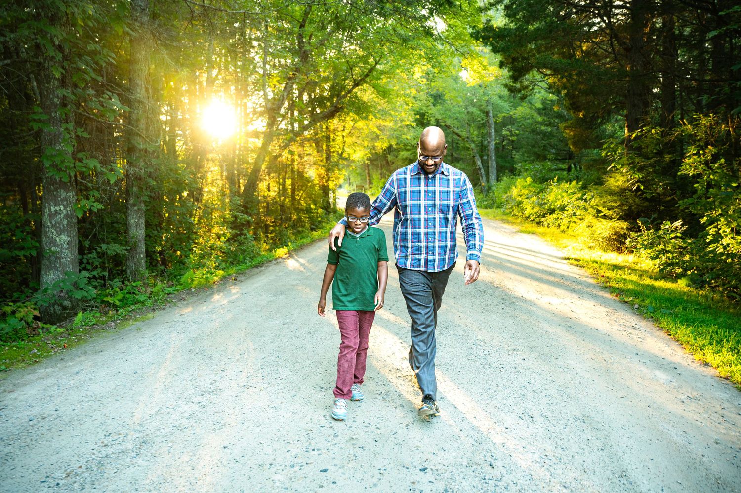 Father and son walking together on a scenic forest road surrounded by lush green trees with warm sunlight filtering through.