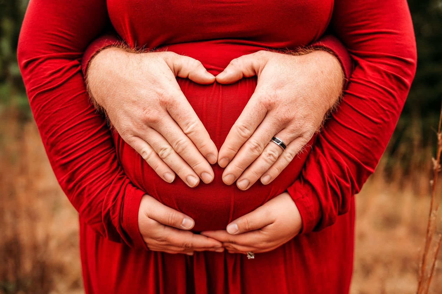 Couple forming heart shape with hands on pregnant belly, woman wearing red maternity dress outdoors.