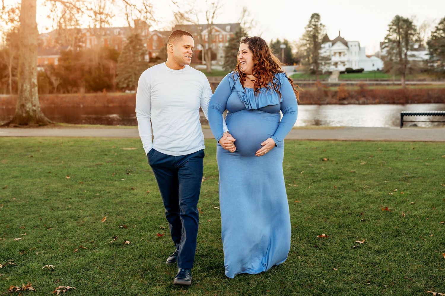 Pregnant couple walking in a park, woman in blue maternity gown holding belly, man in white shirt and jeans smiling at her.