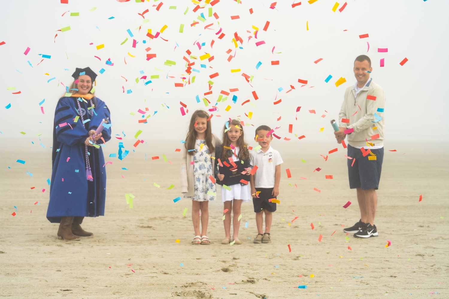 Graduate in blue cap and gown celebrates with family on foggy beach as colorful confetti falls around them.