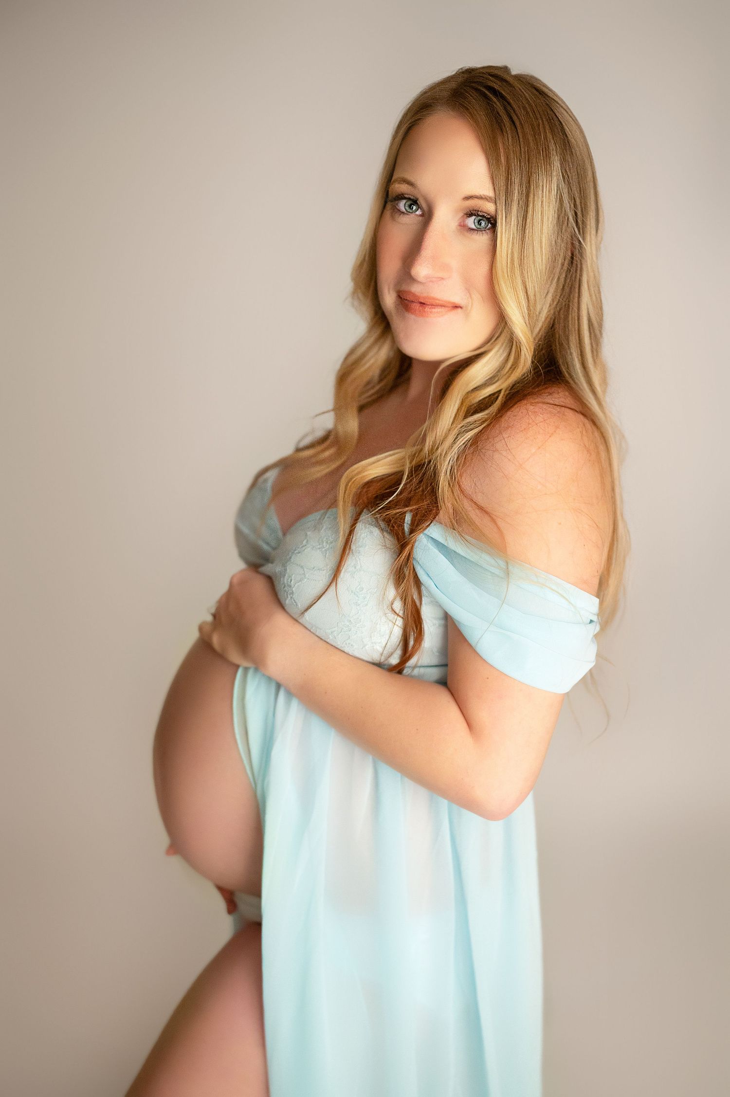 Pregnant woman in light blue off-shoulder maternity gown cradling baby bump in a studio portrait.