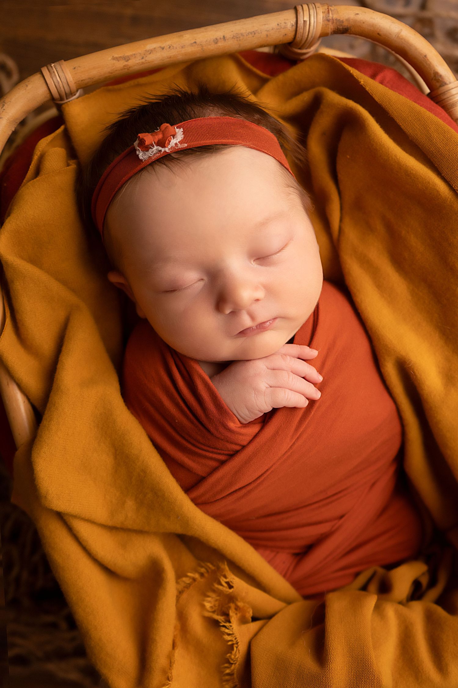 Sleeping newborn baby swaddled in rust orange wrap with matching headband, nestled in a wicker basket with golden blanket.