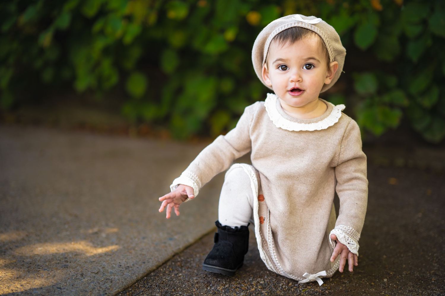 Adorable baby girl in beige knit romper and hat sitting on pavement with green foliage background.
