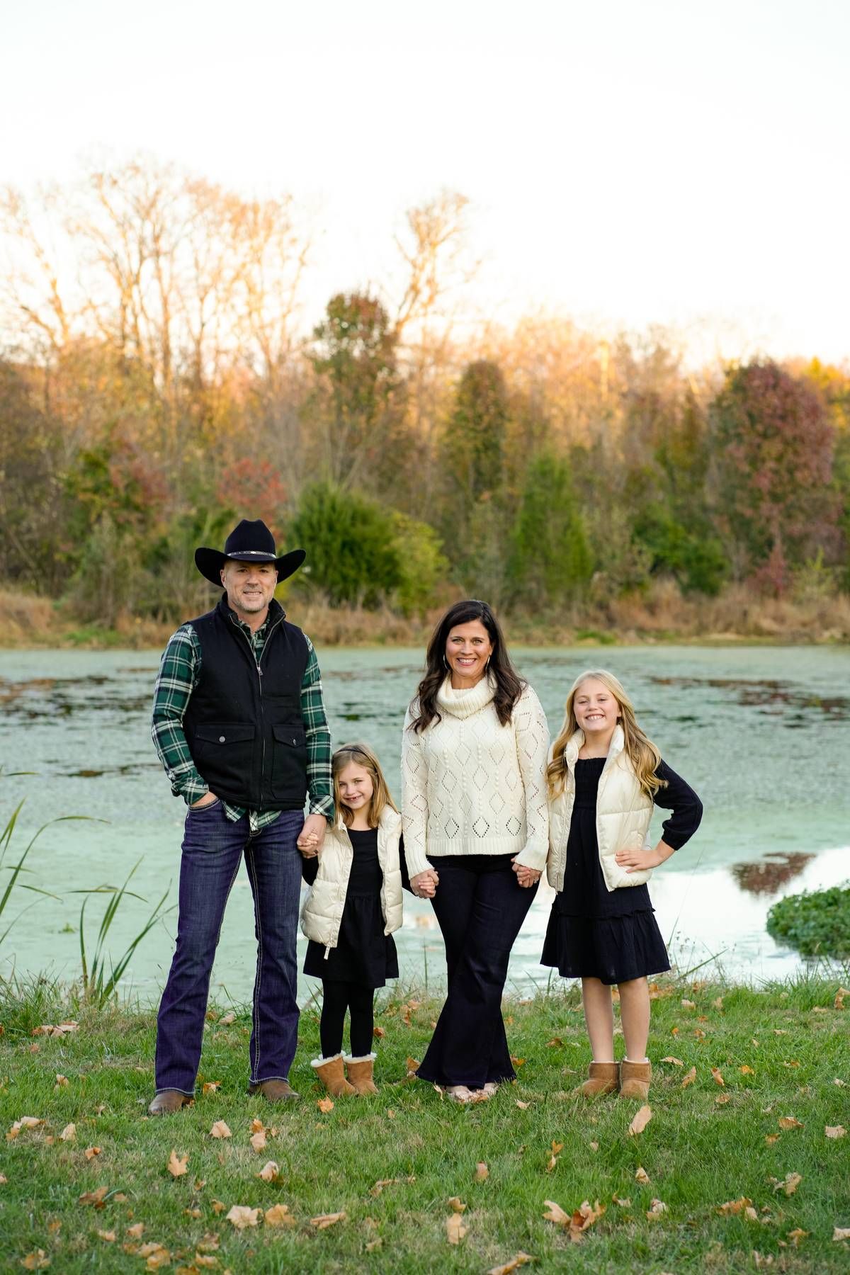 A family of 4 standing holding hands in front of a pond in the fall season, during their family photo session