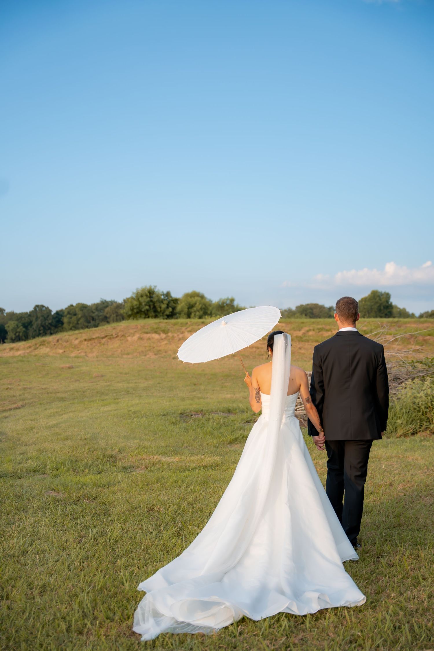 a newlywed couple walking in a grassy field while holding a parasole umbrella