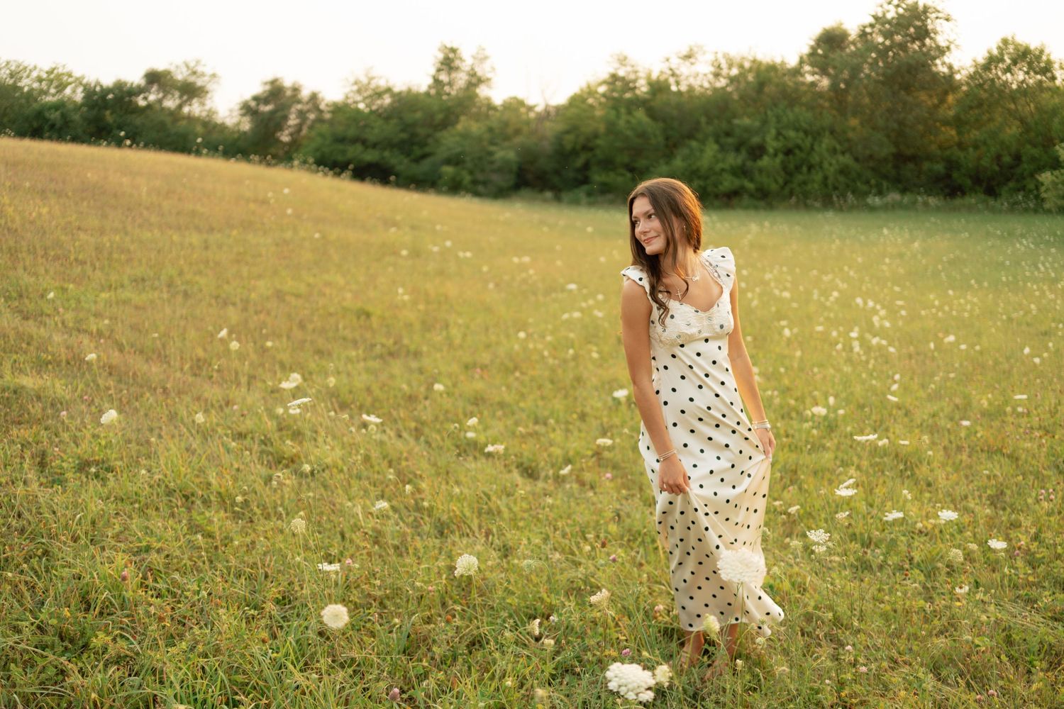 a high school senior girl in a white dress walking in a green grassy field smiling during her senior photo session.