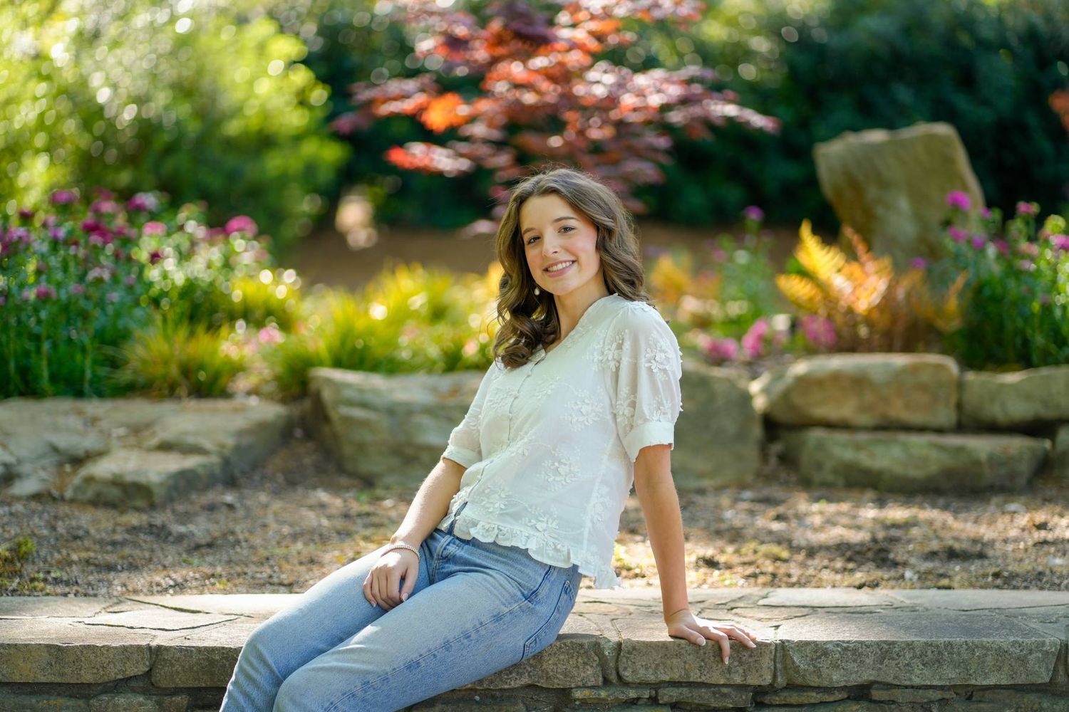 high school girl senior wearing jeans and a white shirt sitting on a rock bench with greenery behind her during her seniro photo session.