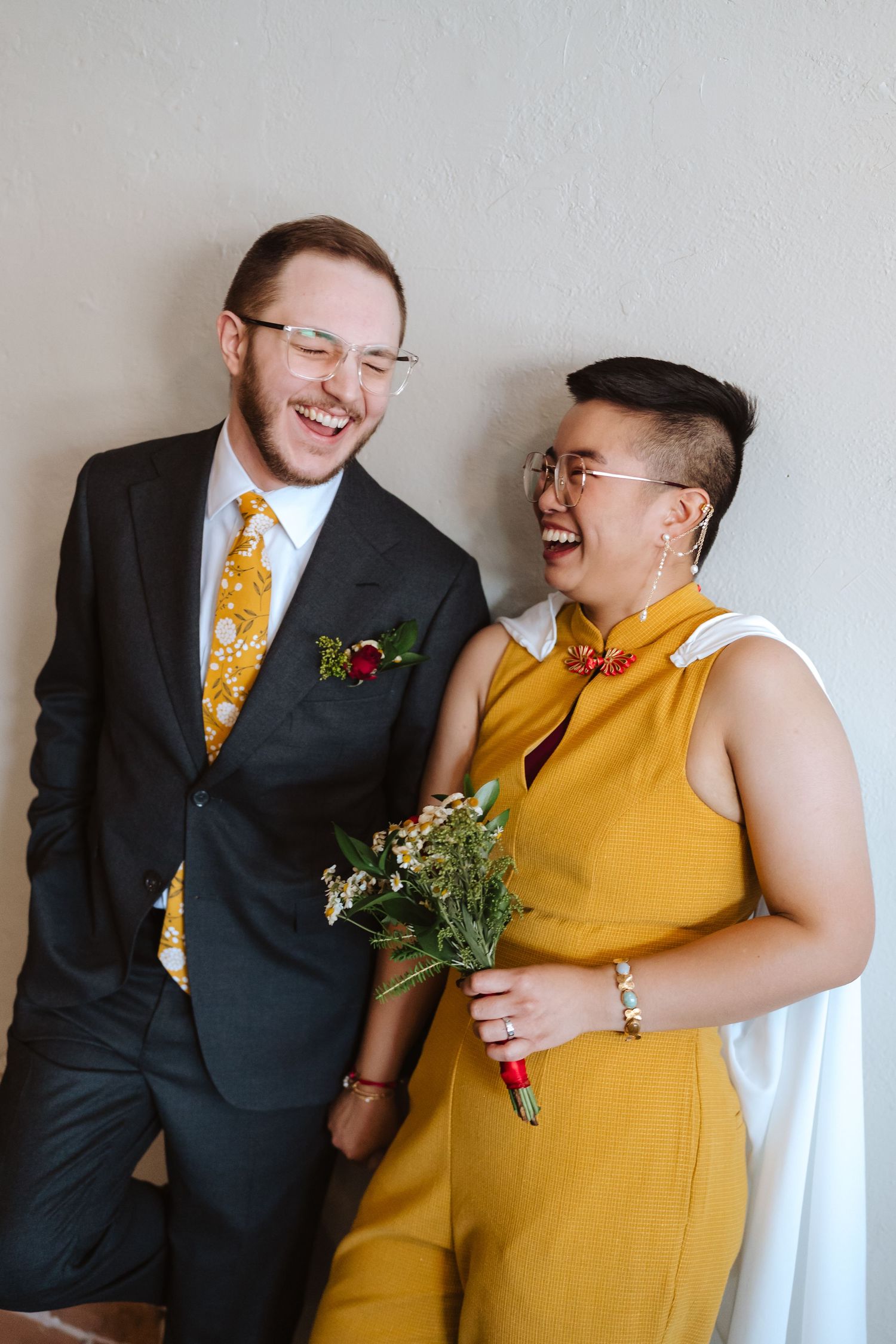 Laughing couple in wedding attire, one in a dark suit with a yellow tie and boutonniere, the other in a mustard jumpsuit holding a bouquet.