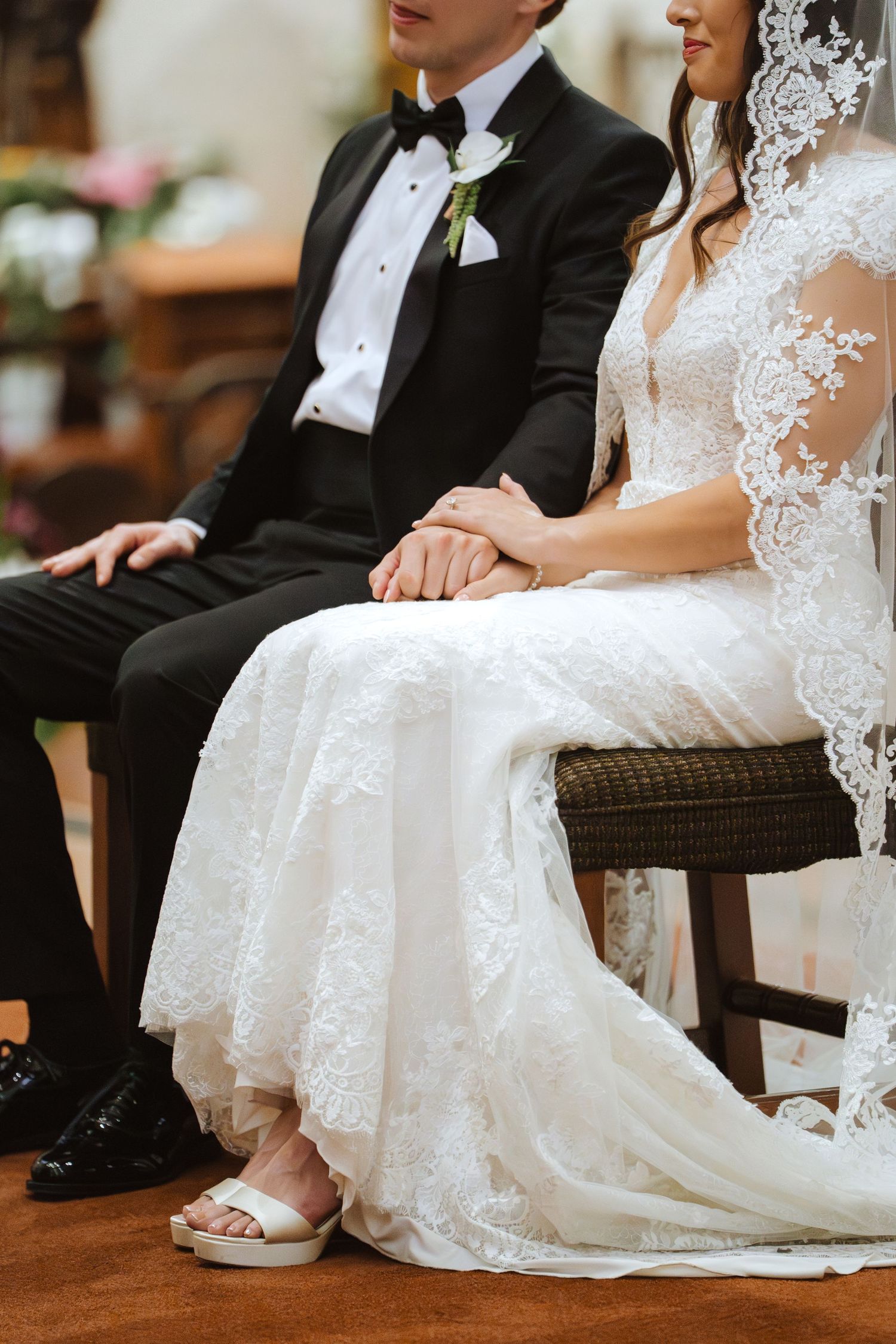 Seated bride in a lace wedding gown and veil holds hands with a groom in a black tuxedo and bow tie during a Catholic wedding ceremony.