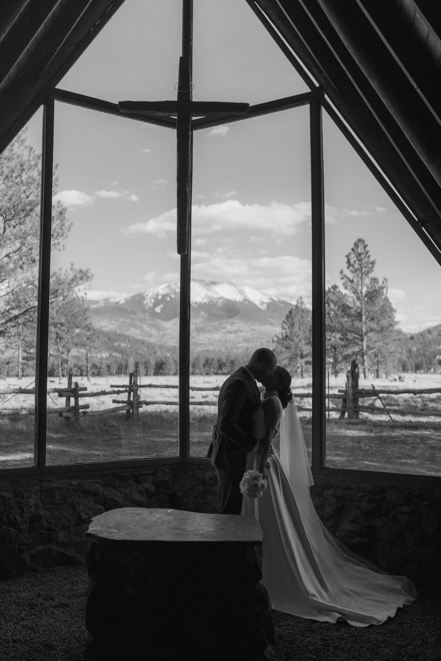 Bride and groom kiss inside a rustic mountain chapel with a large cross-shaped window framing snowy peaks, pine trees, and a wooden fence.