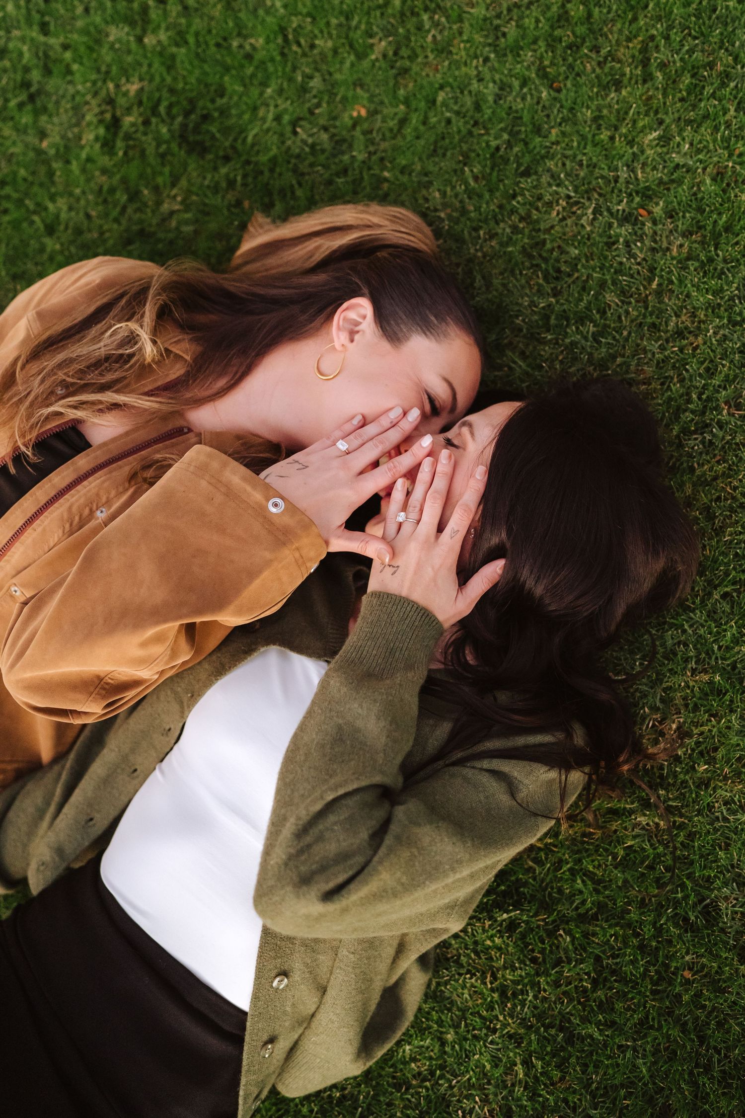 Two women lie on green grass, seen from above, faces close together as they share an intimate moment with their engagement rings.