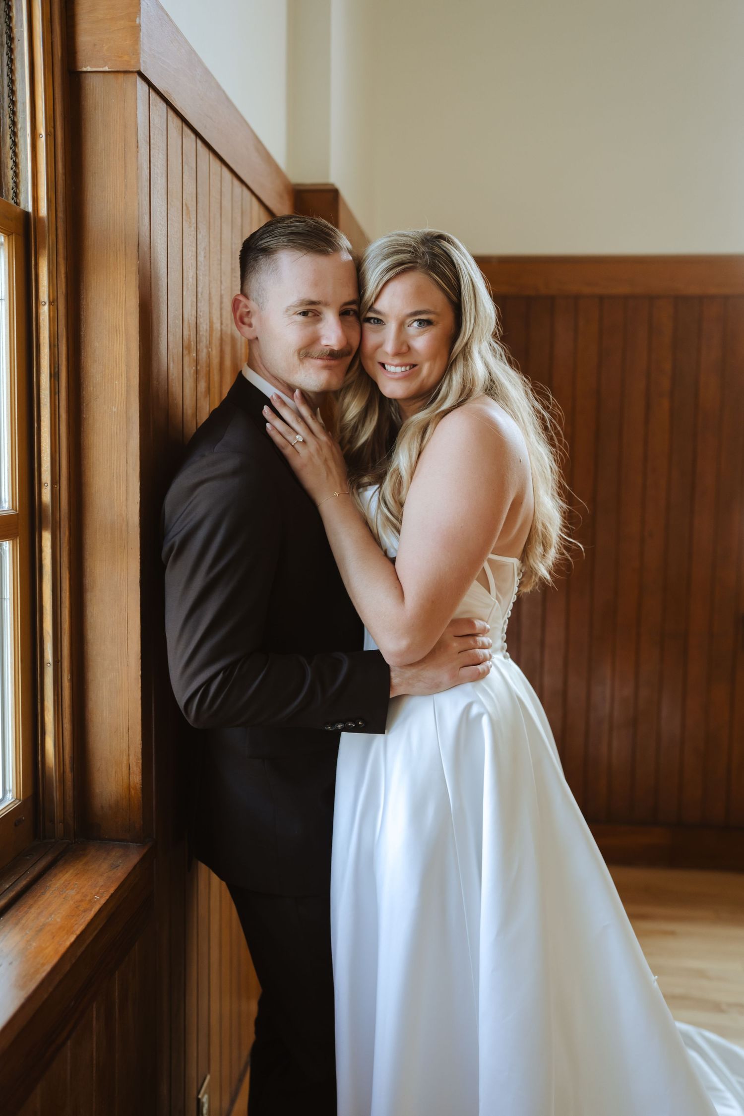 Bride in a white satin gown embraces her groom in a dark suit beside a wood-paneled wall and sunlit window in Prescott Arizona.