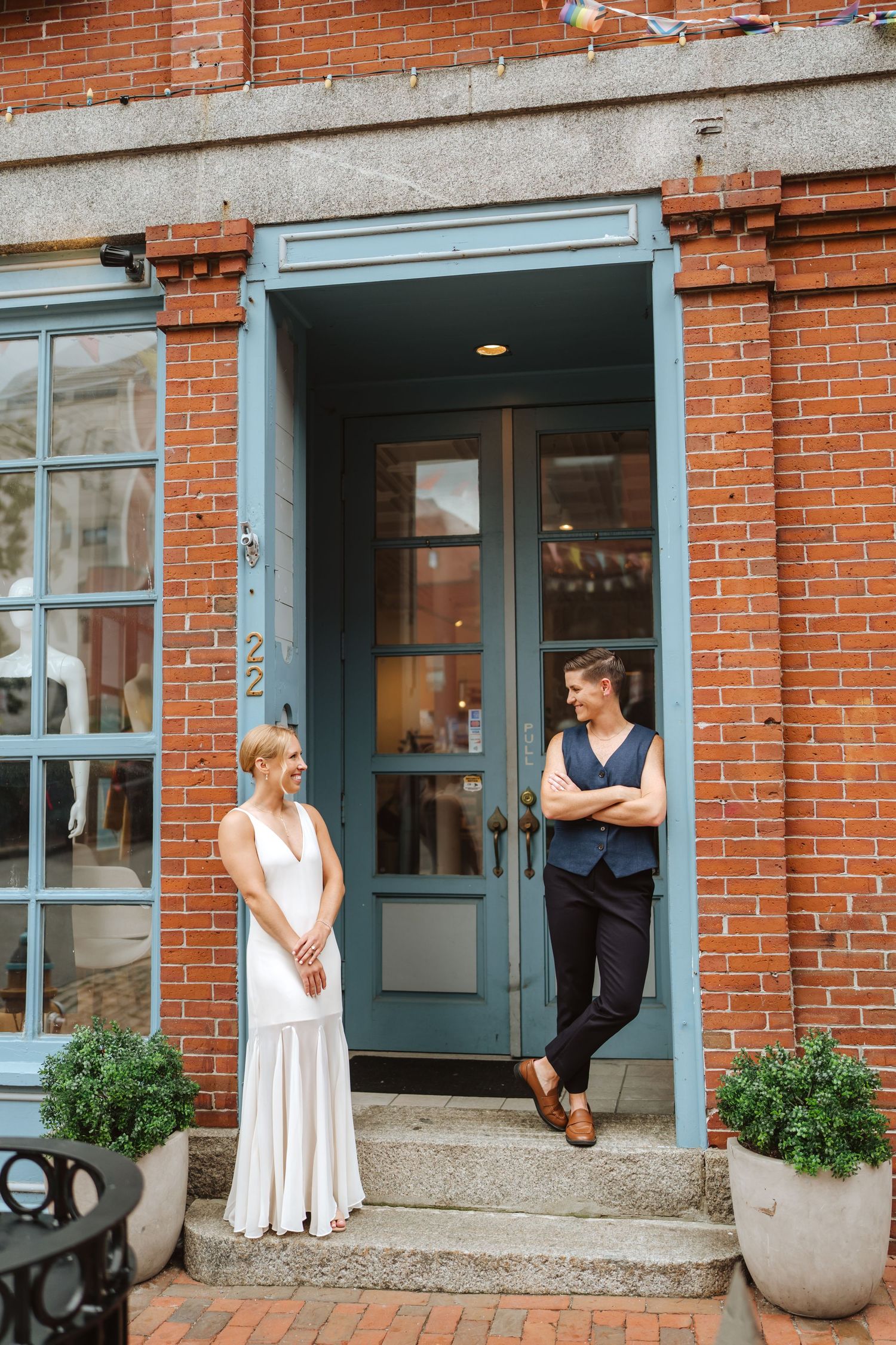 Bride in a white dress and bride in a navy vest lean against a bright blue doorway of a red brick building in Portland Maine.