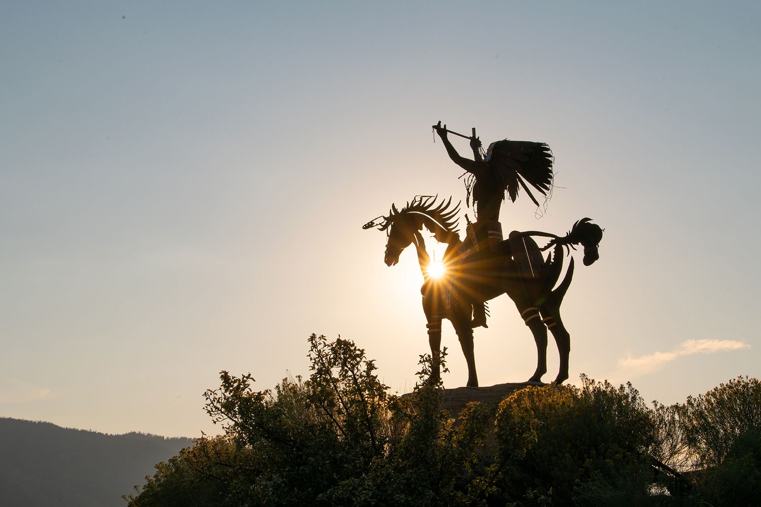 Silhouette of a Native American warrior on horseback statue against a golden sunset sky with mountain backdrop.