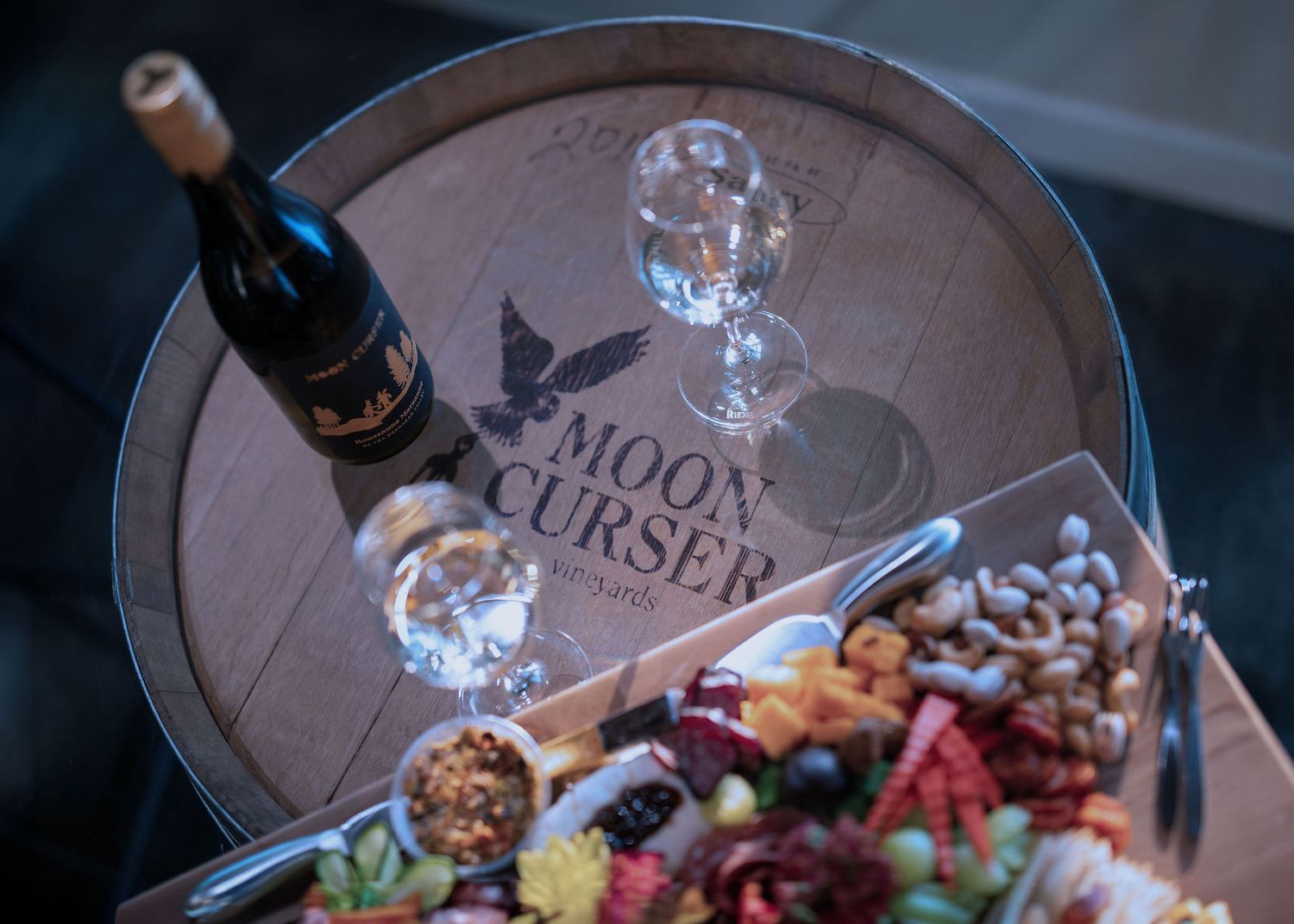 Moon Curser winery barrel display with wine bottle, glasses, and gourmet charcuterie board featuring nuts, fruits, and cheeses.