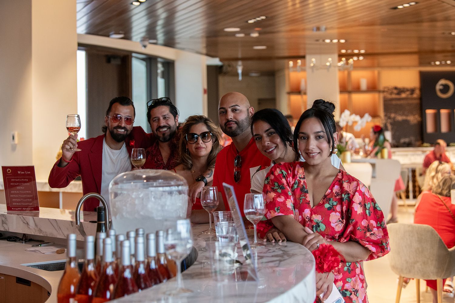 Group of friends smiling and toasting with wine glasses at a stylish bar with rose wine bottles on display.