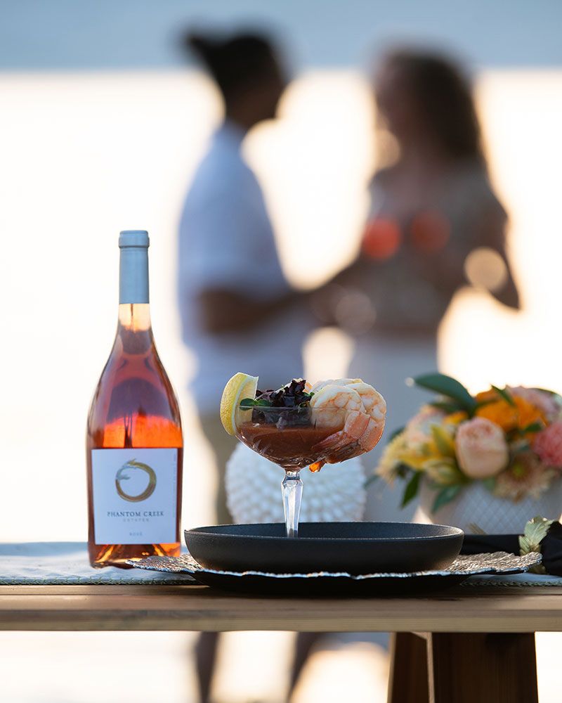 Rose wine bottle and shrimp cocktail on table at sunset with couple in background.