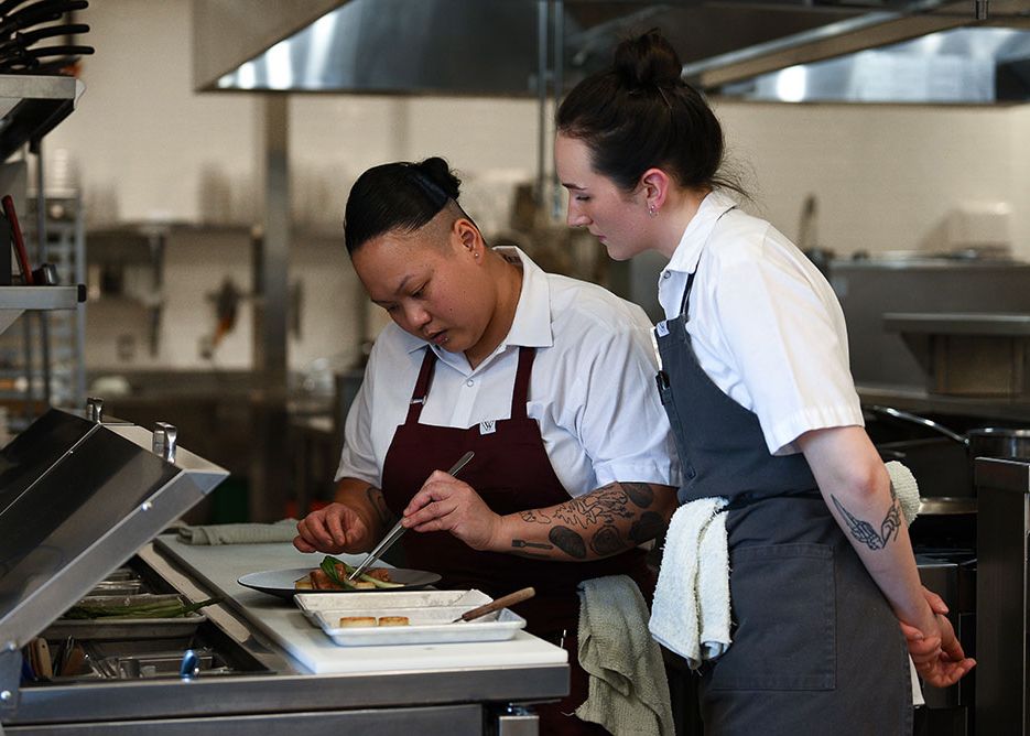 Two chefs in aprons collaborate on plating a dish in a professional restaurant kitchen.
