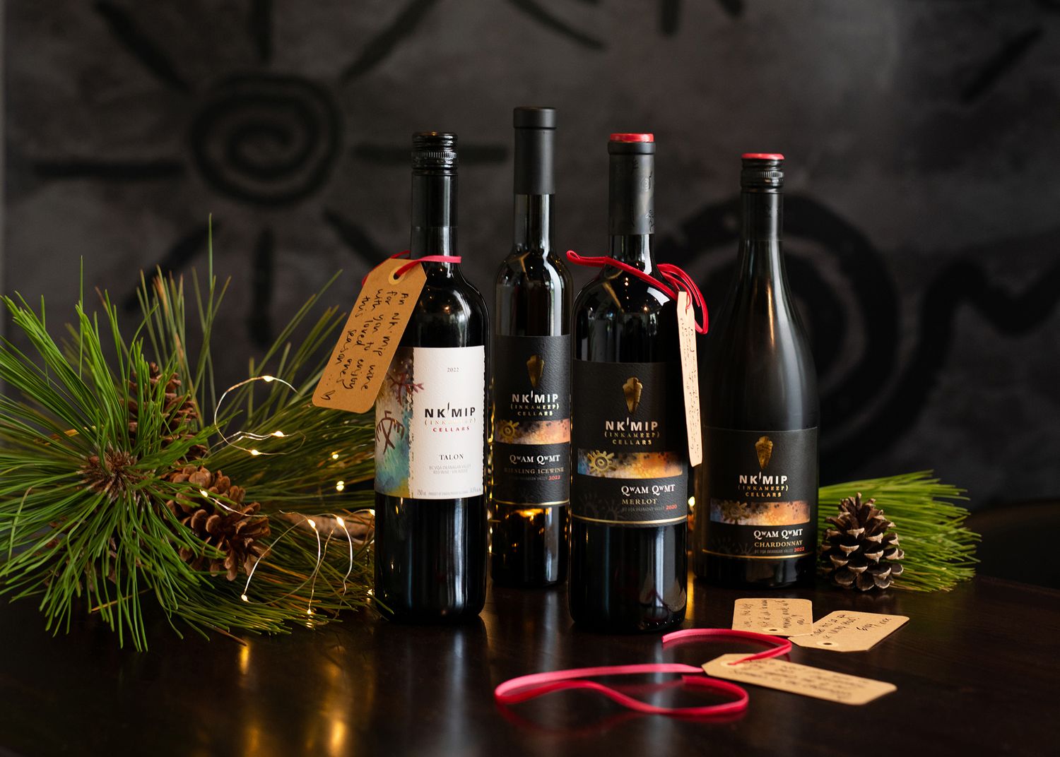 Three bottles of NYW wine with gift tags displayed among pine branches and pinecones on a dark surface.