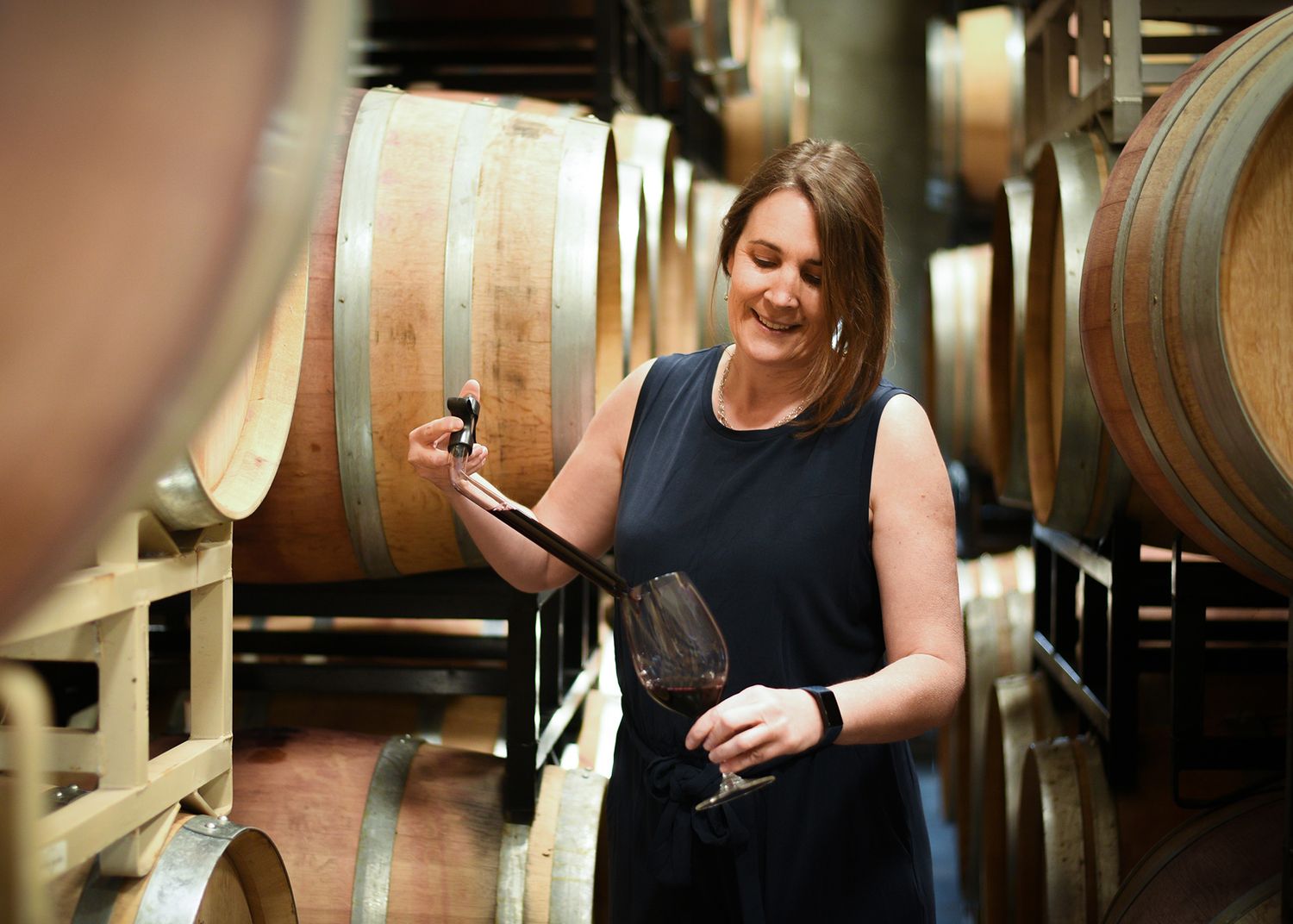 Woman examining red wine from barrel in a winery cellar surrounded by oak barrels.