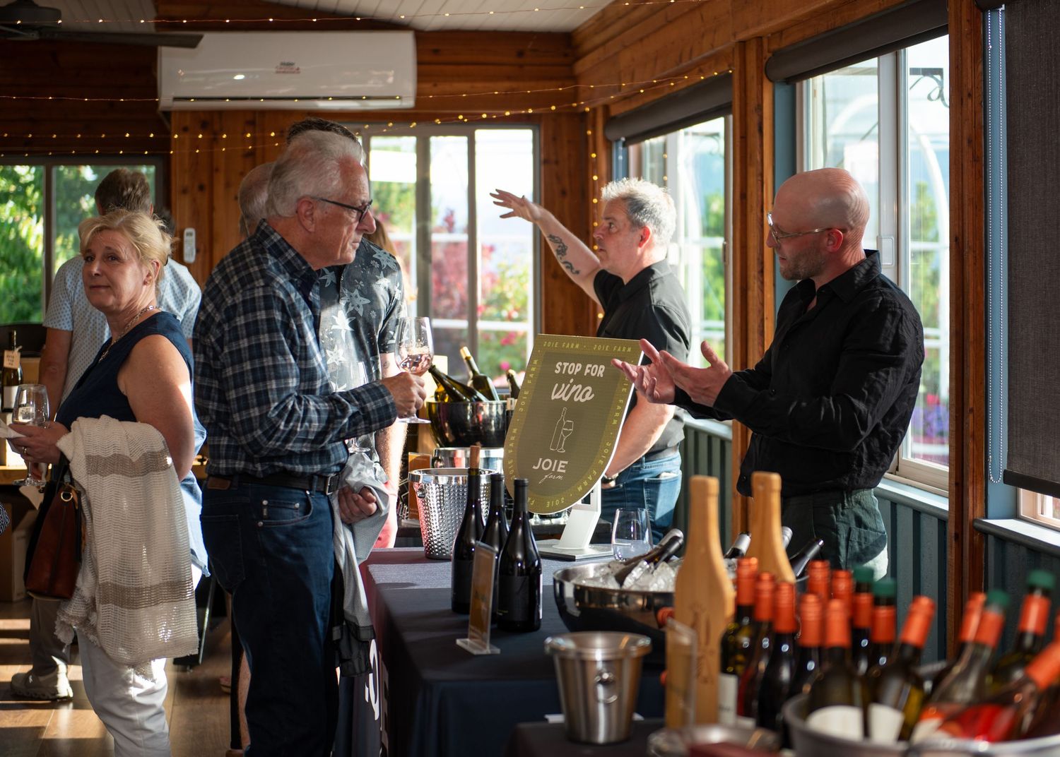 Guests enjoying a wine tasting event indoors, with bottles displayed on a table and a man gesturing enthusiastically.