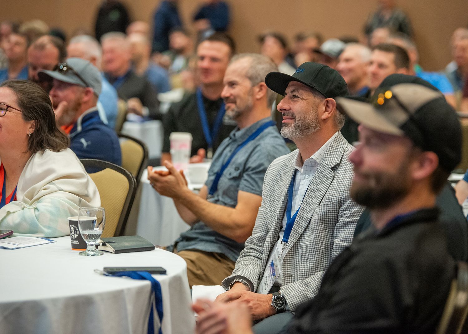 Audience, including man with black ball cap and grey blazer, listening to conference speaker.