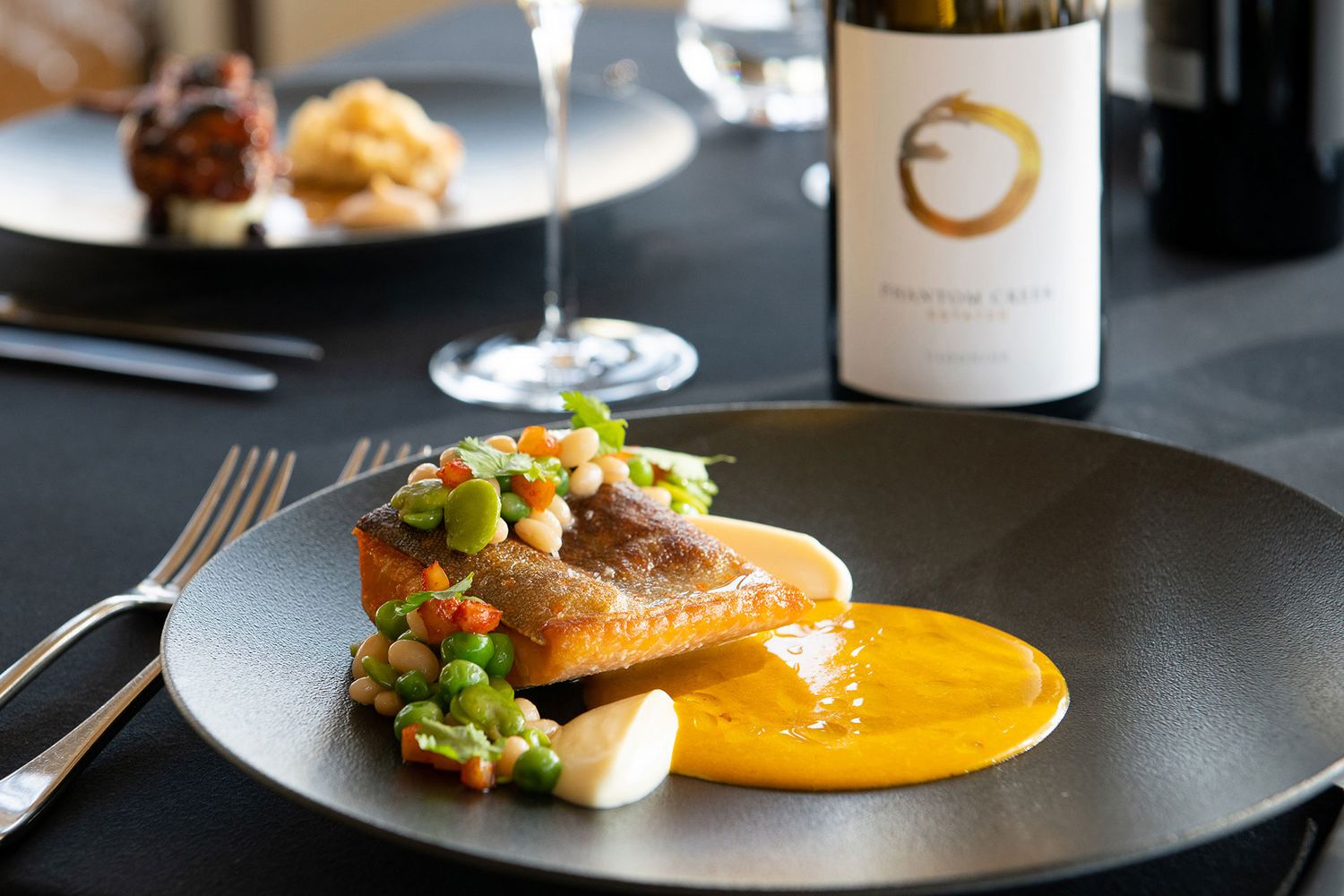 Plated gourmet dish featuring seared fish over orange purée with vegetables, served with a glass of wine and bottle in the background.