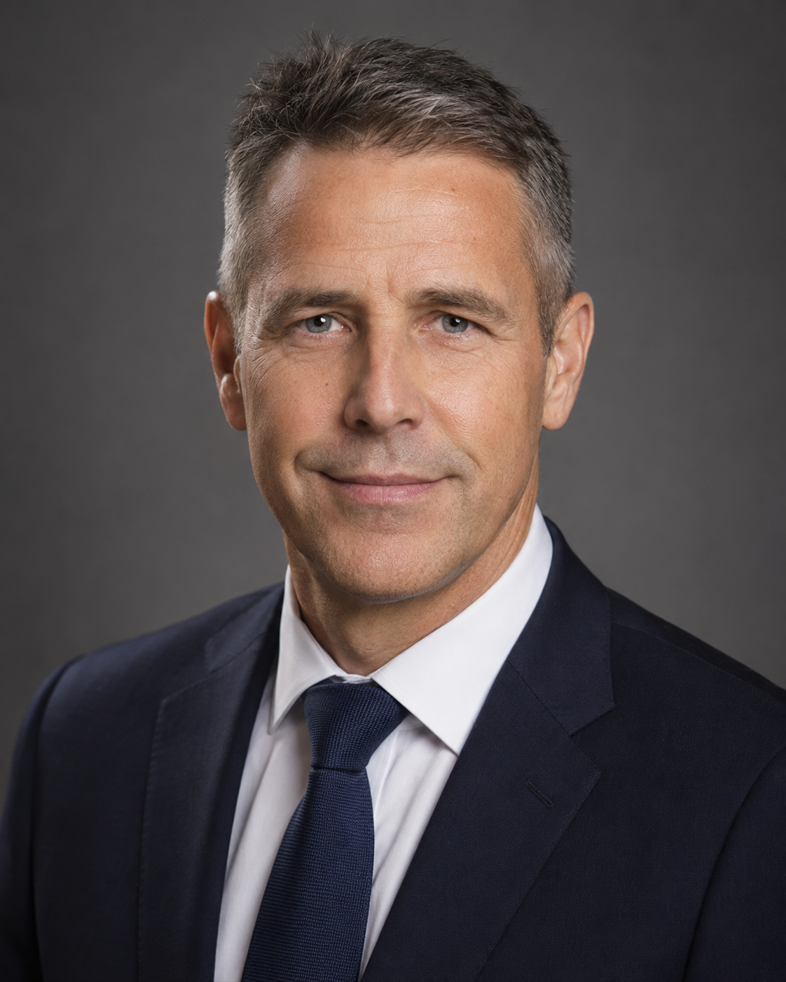 Professional headshot of a middle-aged man with gray hair wearing a navy suit and tie against a gray background.