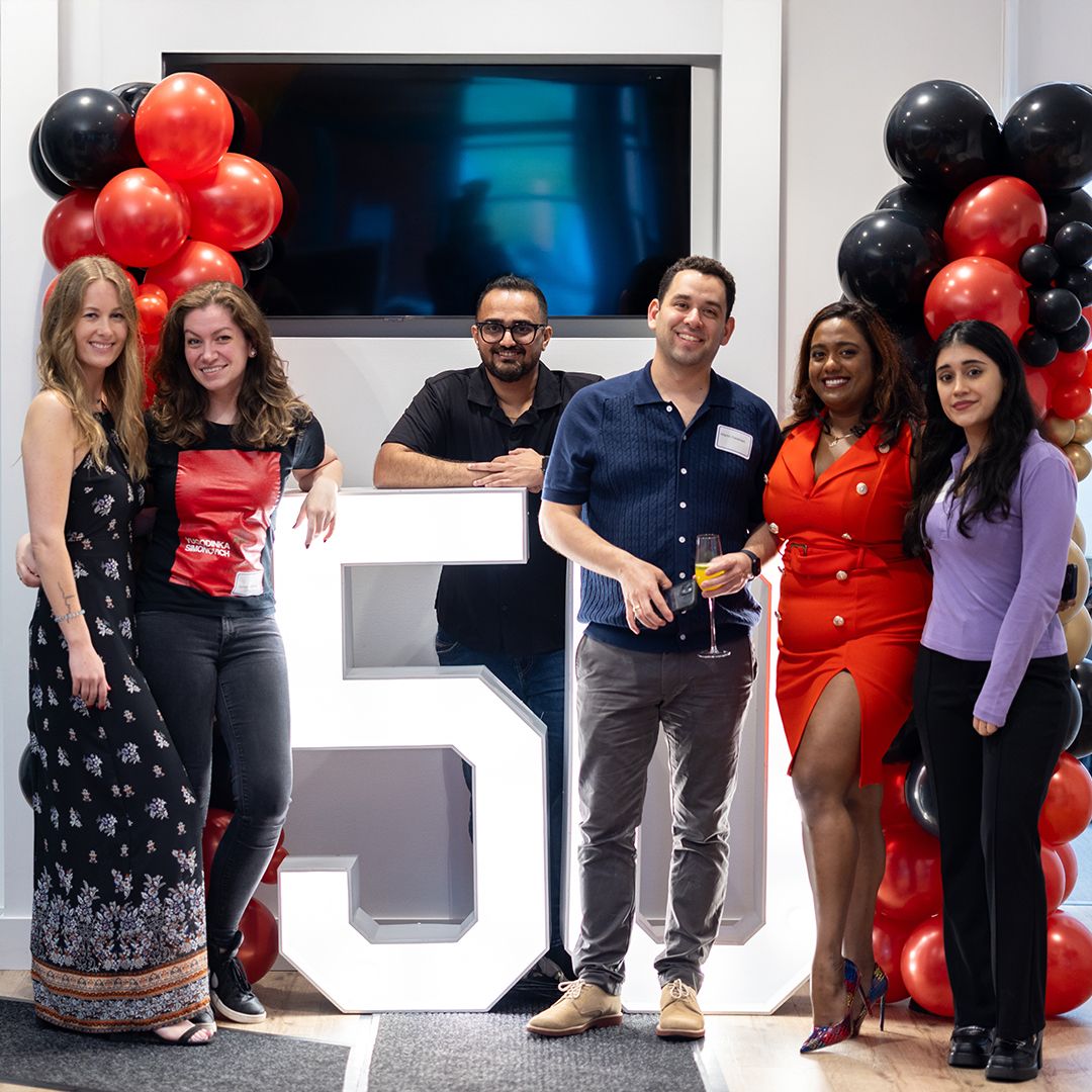 Group of six colleagues posing beside a large number 5 with red and black balloon decorations at a company anniversary celebration.