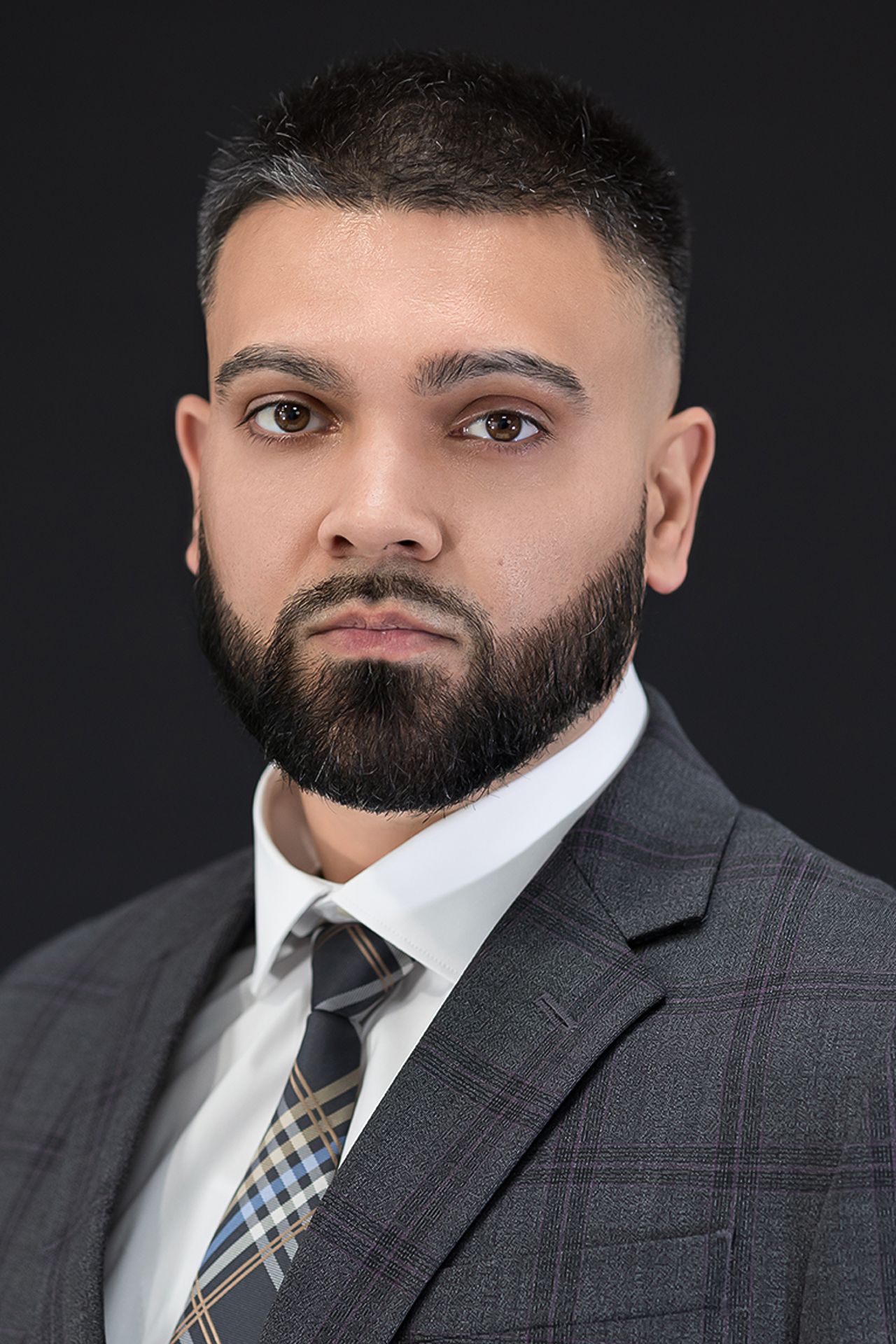 Professional headshot of a bearded man in a gray plaid suit and plaid tie against a dark background.
