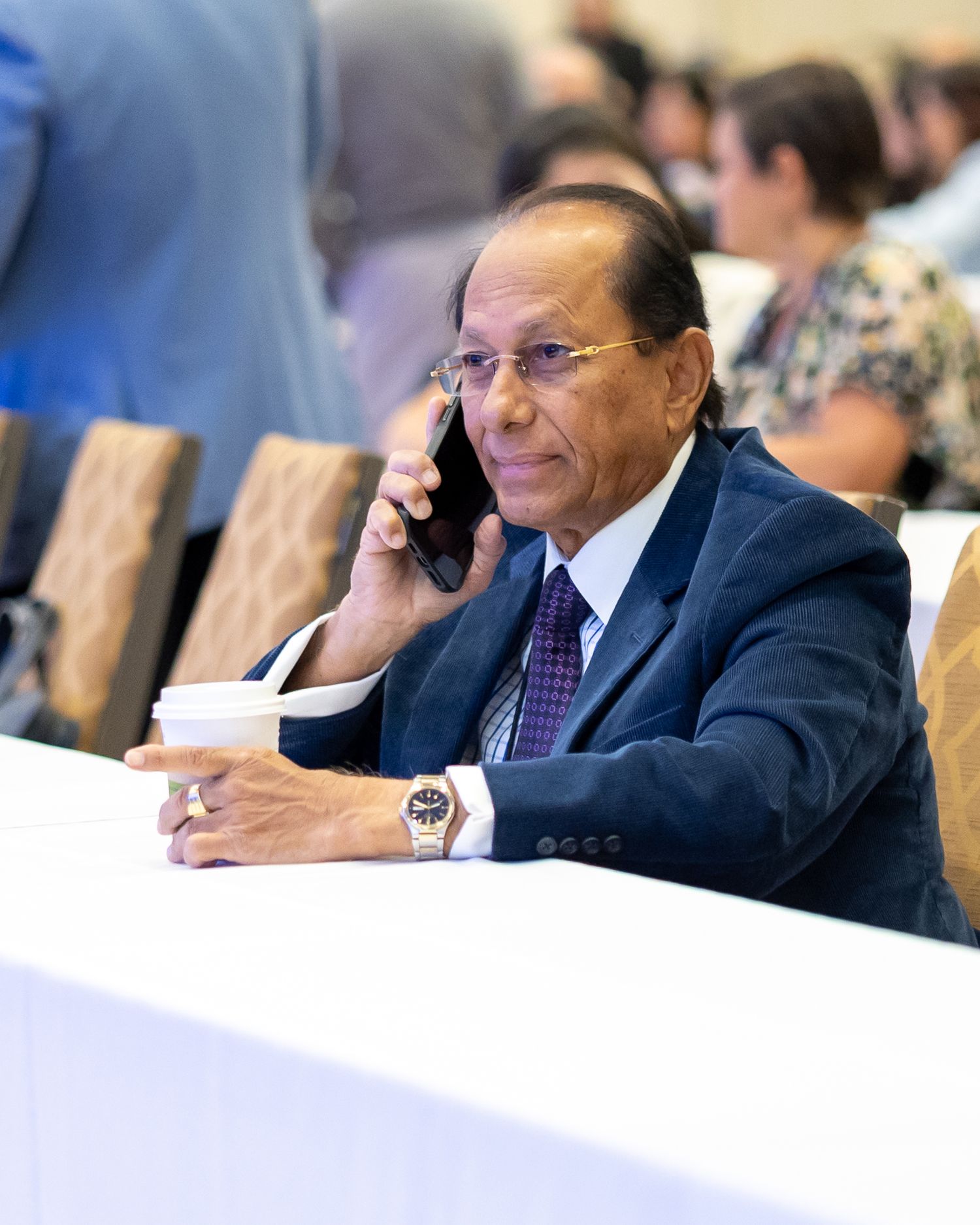 Elderly man in a navy suit and purple tie talking on a phone while holding a coffee cup at a formal event.